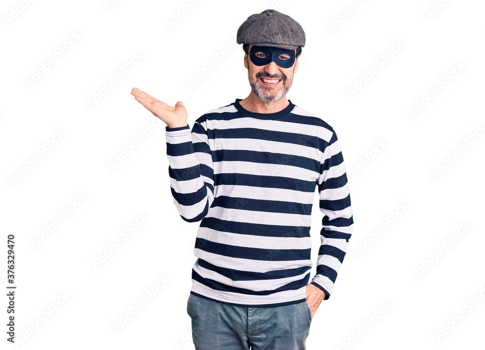 Middle age handsome man wearing burglar mask smiling cheerful presenting and pointing with palm of hand looking at the camera. Middle age handsome man wearing burglar mask smiling cheerful presenting and pointing with palm of hand looking at the camera.