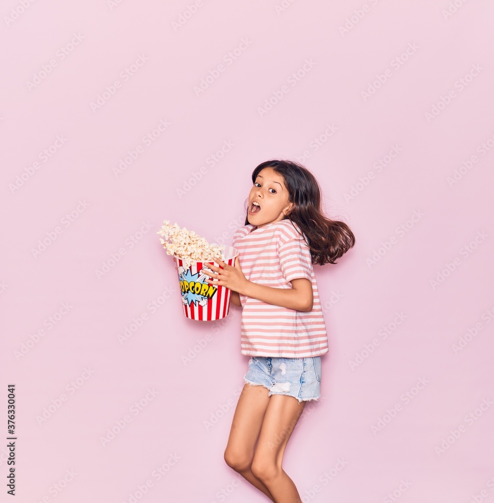 Adorable hispanic child girl surprised with open mouth holding bucket of popcorn jumping over isolated pink background Adorable hispanic child girl surprised with open mouth holding bucket of popcorn jumping over isolated pink background