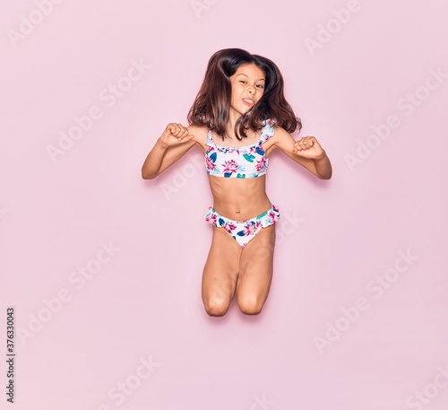 Adorable hispanic child girl on vacation wearing bikini smiling happy. Jumping with smile on face over isolated pink background