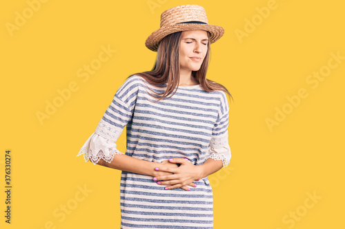 Beautiful caucasian woman wearing summer hat with hand on stomach because indigestion, painful illness feeling unwell. ache concept.
