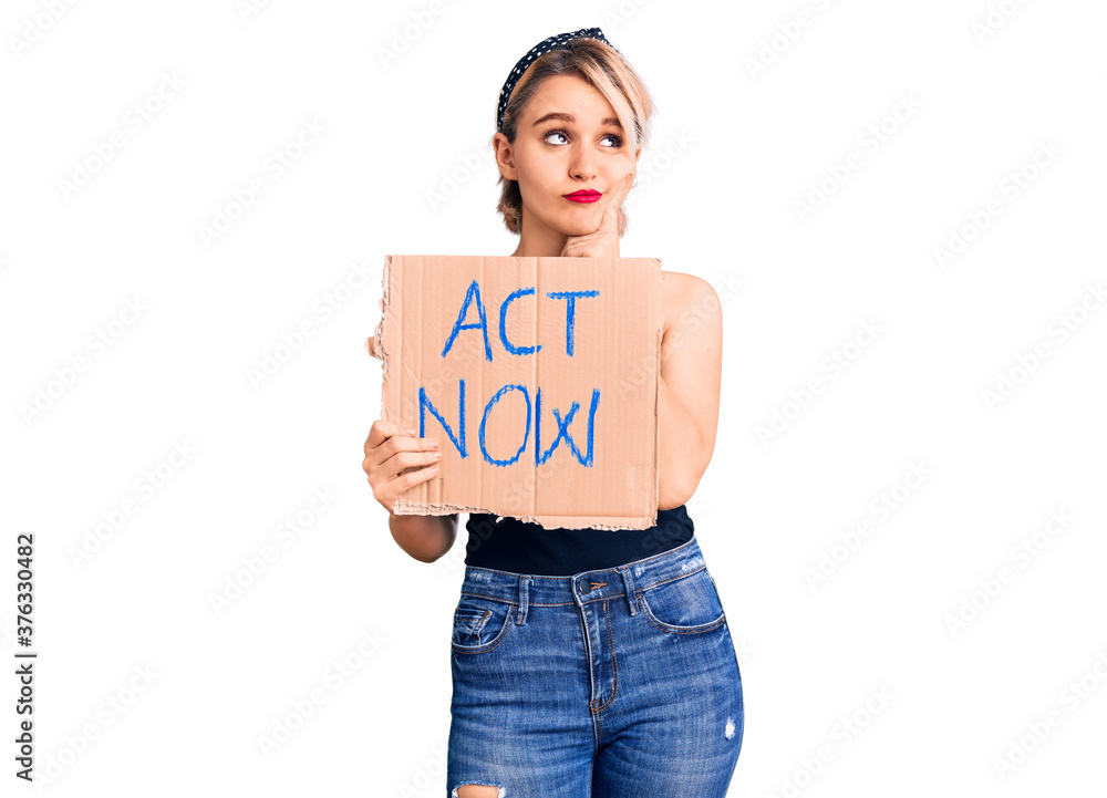 Young beautiful blonde woman holding act now banner serious face thinking about question with hand on chin, thoughtful about confusing idea Young beautiful blonde woman holding act now banner serious face thinking about question with hand on chin, thoughtful about confusing idea