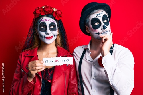 Couple wearing day of the dead costume holding trick or treat paper serious face thinking about question with hand on chin, thoughtful about confusing idea