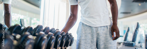 African American man picking up a dumbbell from the rack and looking outside at gym. Male weight training person holding sport equipment with serous look in fitness center photo