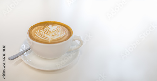 Closeup of white cup of hot coffee latte with milk foam heart shape art on plate and spoon on white table with copy space.