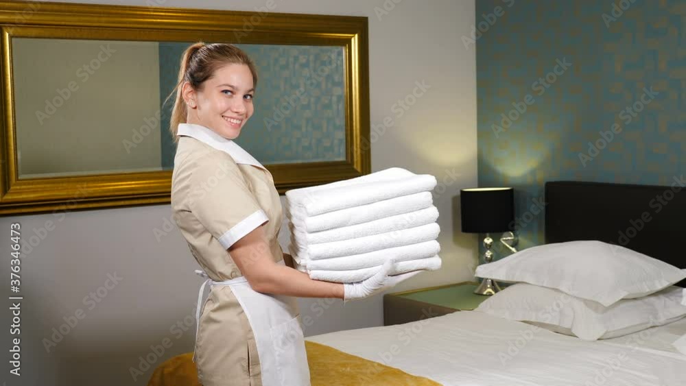 Professional cleaning service. Pretty maid holding stack of folded clean towels then throwing it up smiling at camera with king-sized bed in background. Room preparation for guests. 4 k video Professional cleaning service. Pretty maid holding stack of folded clean towels then throwing it up smiling at camera with king-sized bed in background. Room preparation for guests. 4 k video