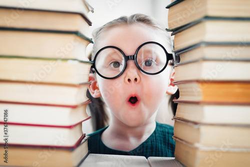 Shocked little blonde kid girl 4-6 years old in library among many books. Back to school concept.