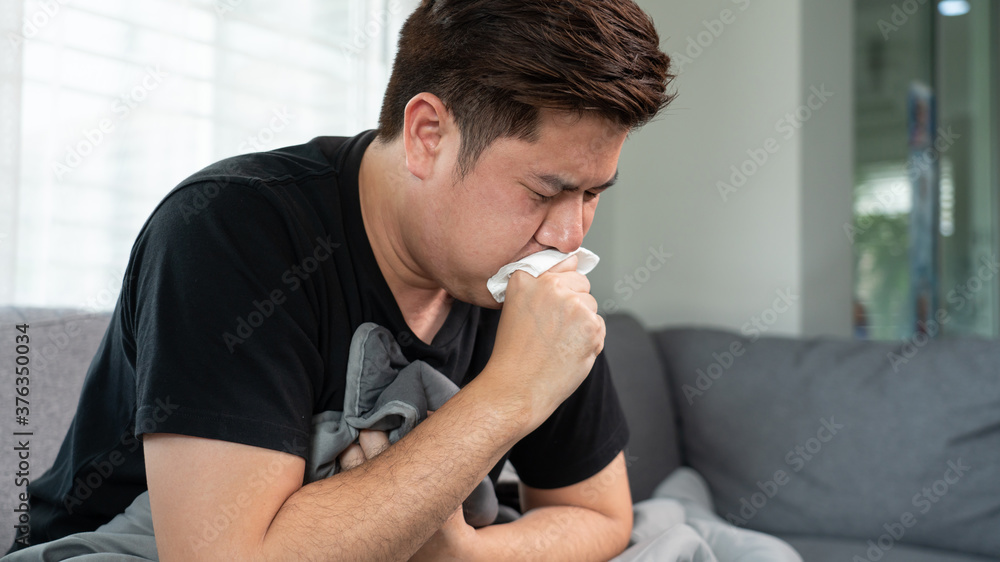 Asian man with seasonal infections Cold Blowing His Nose and sneezing into Tissue with headache lying on sofa with high fever and a flu Asian man with seasonal infections Cold Blowing His Nose and sneezing into Tissue with headache lying on sofa with high fever and a flu