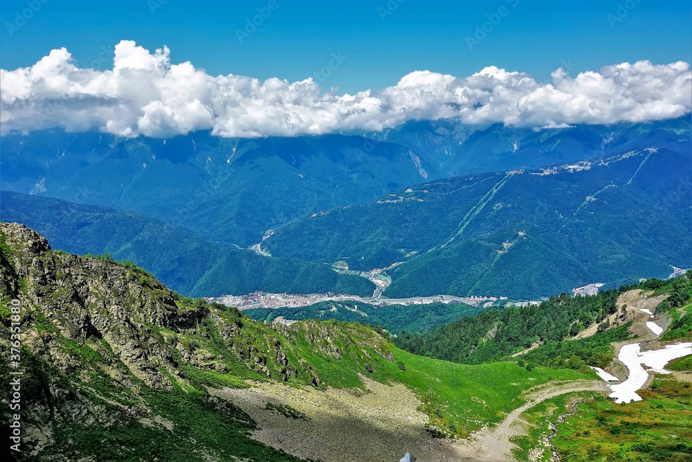 Obraz premium Alpine landscape. In the valley there is green grass, islands of snow. A village is visible below. The peaks of the mountain range are hidden in the clouds. Summer. Caucasus. Russia.