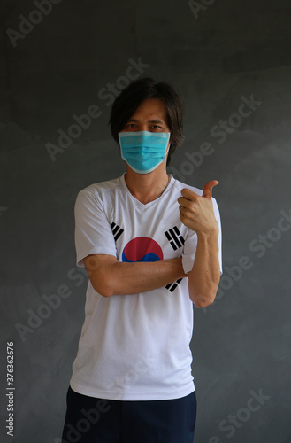 Masked man wearing South Korea flag color of shirt and cross one s arm with thumbs up on dark wall background. photo