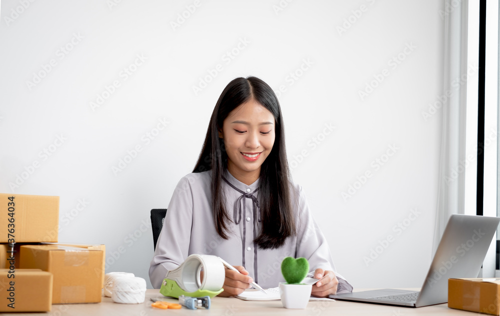 Young Asian woman business owner with many parcel boxes on the table happy online sales job, use your laptop, get an order from customers, take notes, and make arrangements for delivery by post Young Asian woman business owner with many parcel boxes on the table happy online sales job, use your laptop, get an order from customers, take notes, and make arrangements for delivery by post