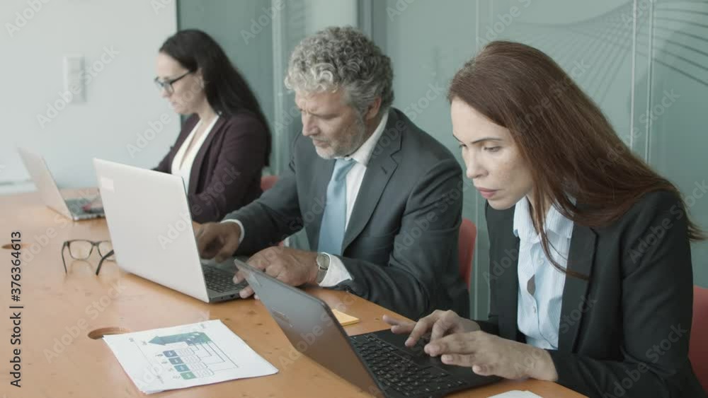 Businesspeople sitting together at desk and working at computers, using laptops in conference room. Medium shot. Communication or wireless technology concept Businesspeople sitting together at desk and working at computers, using laptops in conference room. Medium shot. Communication or wireless technology concept