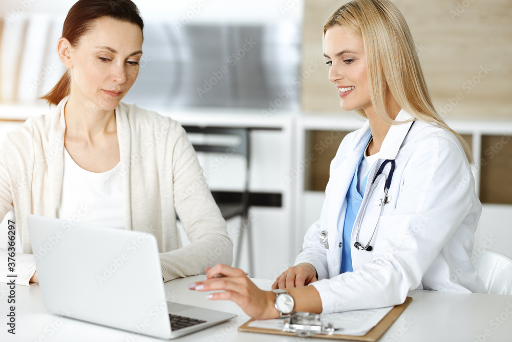Woman-doctor at work in sunny hospital is happy to consult female patient. Blonde physician checks medical history record and exam results while using clipboard Woman-doctor at work in sunny hospital is happy to consult female patient. Blonde physician checks medical history record and exam results while using clipboard