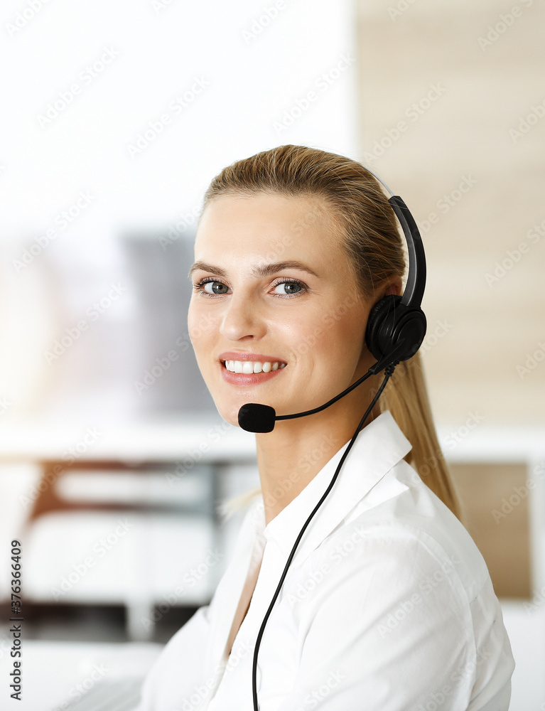 Blond female customer service representative is consulting clients online using headset in sunny office. Call center concept Blond female customer service representative is consulting clients online using headset in sunny office. Call center concept