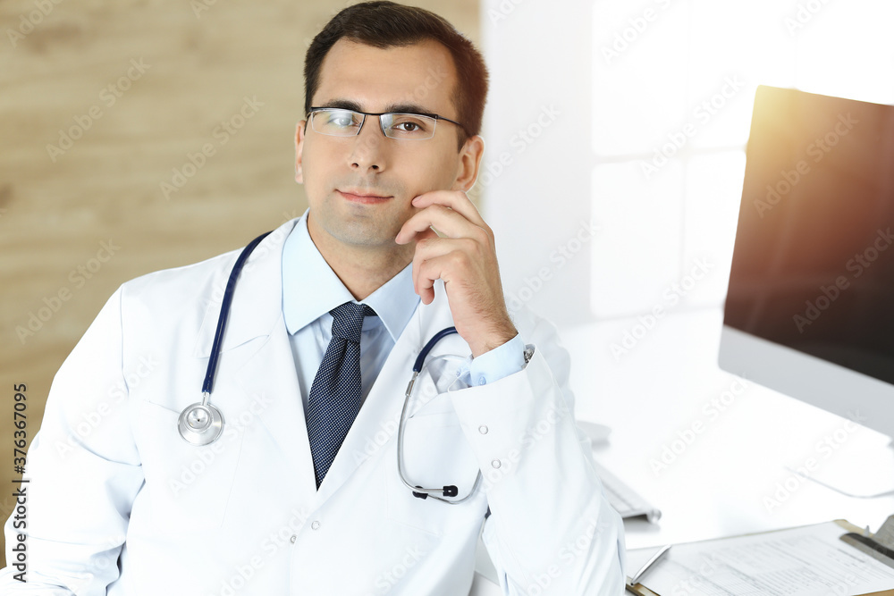 Man-doctor sitting at the desk at his working place and smiling. Perfect medical service in sunny clinic Man-doctor sitting at the desk at his working place and smiling. Perfect medical service in sunny clinic