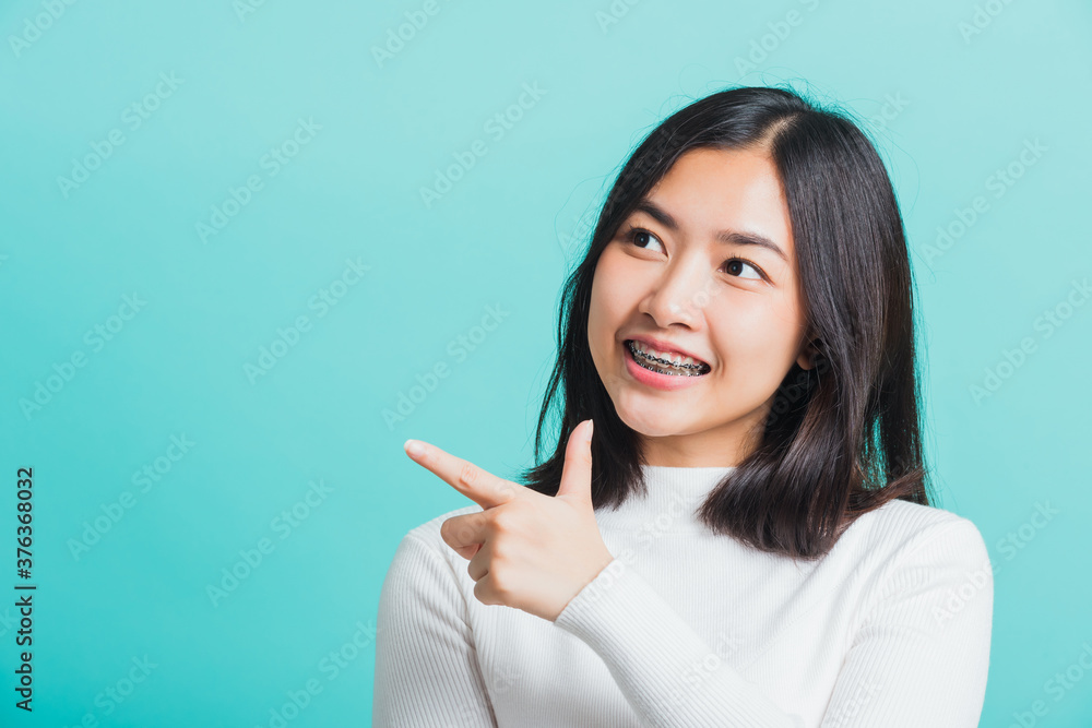 Portrait of Asian teen beautiful young woman smile have dental braces on teeth laughing point finger side away blank copy space, studio shot isolated on blue background, medicine and dentistry concept Portrait of Asian teen beautiful young woman smile have dental braces on teeth laughing point finger side away blank copy space, studio shot isolated on blue background, medicine and dentistry concept
