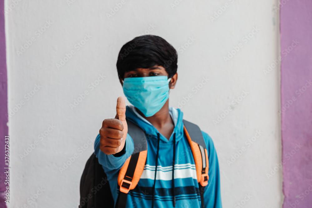 Portrait of an Indian school kid wearing medical mask giving thumbs up in front of the camera, New normal concept, Back to school after corona virus pandemic, School safety concept after corona virus