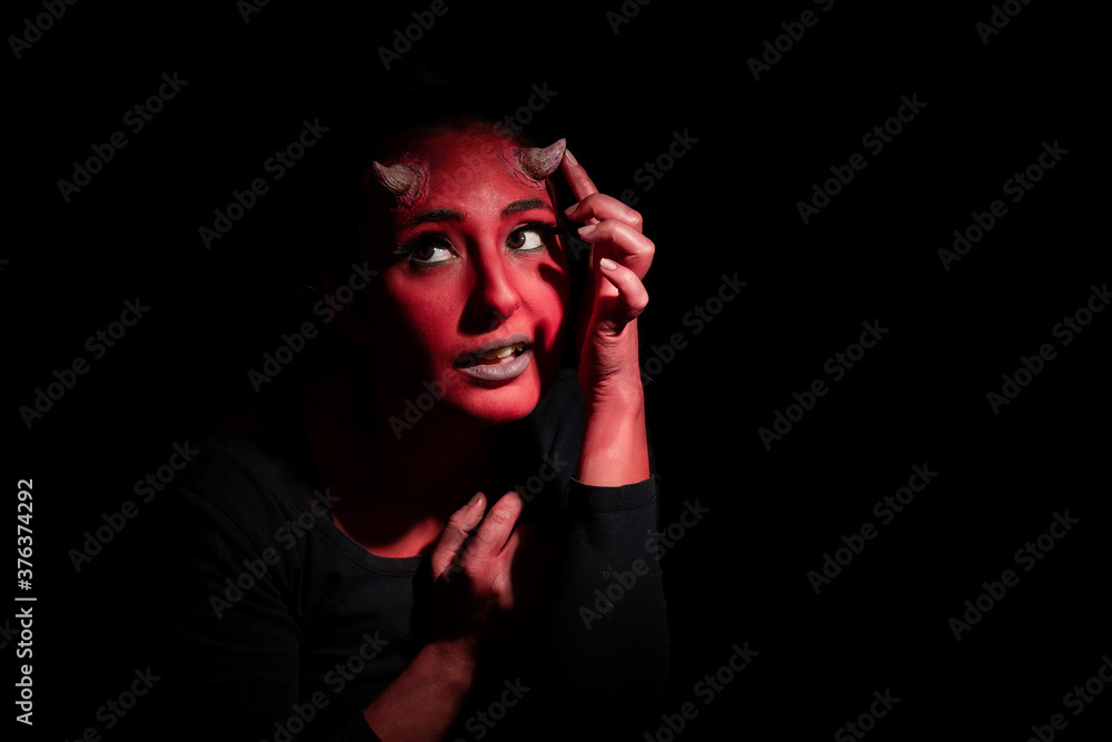 Halloween concept. Headshot of demon woman with red face makeup and small horns, wants to look spooky, poses against black background with copy space. Demon woman with red face touches her small horns