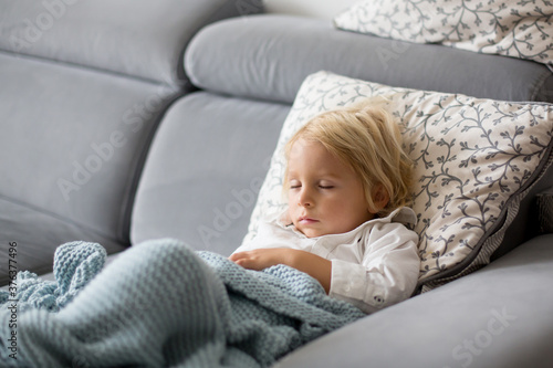 Sick child, toddler boy lying on the couch with his white puppy in living room with fever, resting