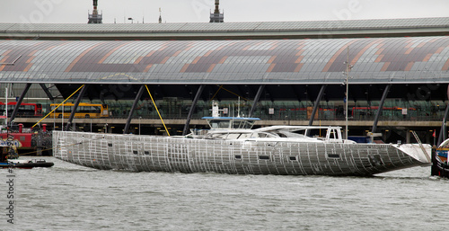 Aluminium hull. Transport of a super yacht hull on a pontoon with tugboats at the river. . Ship building industry. Super sailing yacht. Netherlands. Het IJ. Amsterdam. Central station.