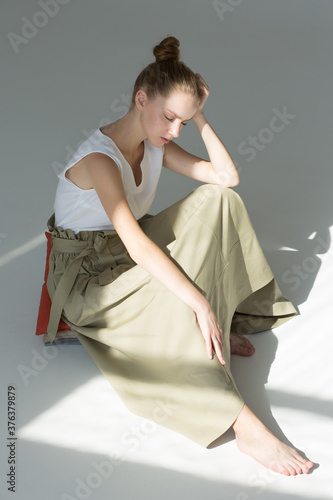 A young woman sits on the floor in the studio. She is wearing a long green skirt and a white summer tank top. The eyes are closed. Natural makeup.