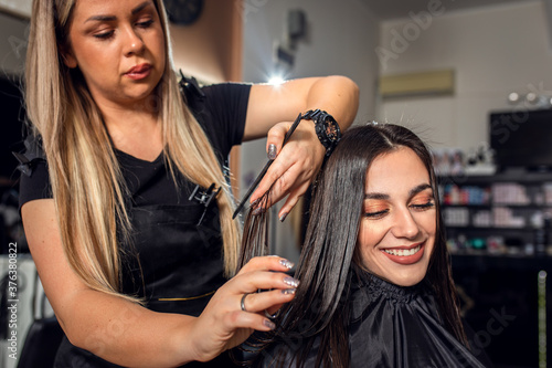 Female hairdresser makes hairstyle on young brunette woman in salon cutting hair with scissors.