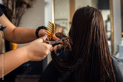 Female hairdresser makes hairstyle on young brunette woman in salon cutting hair with scissors.