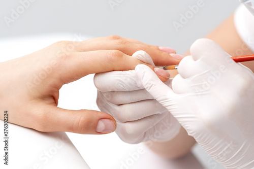 Manicure master in rubber gloves applying transparent nail polish on female nails in beauty salon