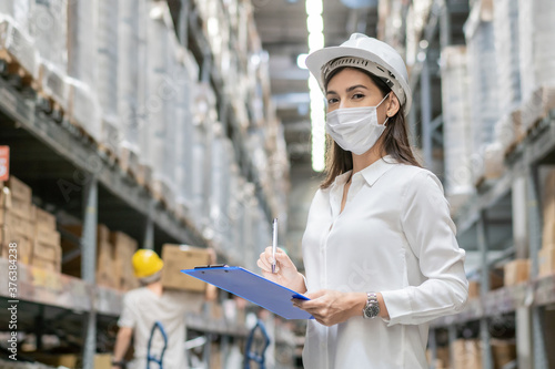 Female construction worker wearing protective face mask writing on clipboard checking product on aisle in warehouse factory. industrial social responsibility prevention Coronavirus. new normal working