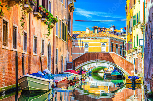 Venice cityscape with gondolas and motor boats moored on narrow water canal near colorful buildings and stone bridge, Veneto Region, Northern Italy, blue sky in summer day, typical venetian view