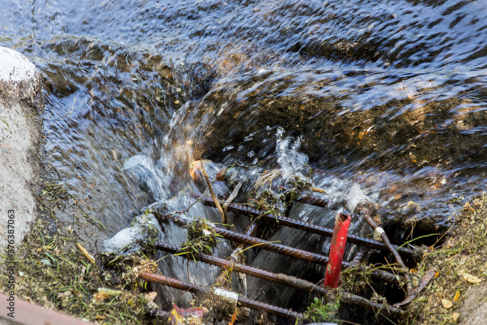 Obraz premium Accident of water supply, sewage. Water drains out of sewer hatch and flows down road. Emergency discharge of sewage into storm drainage system. Water flows on way to the rain storm sewage system