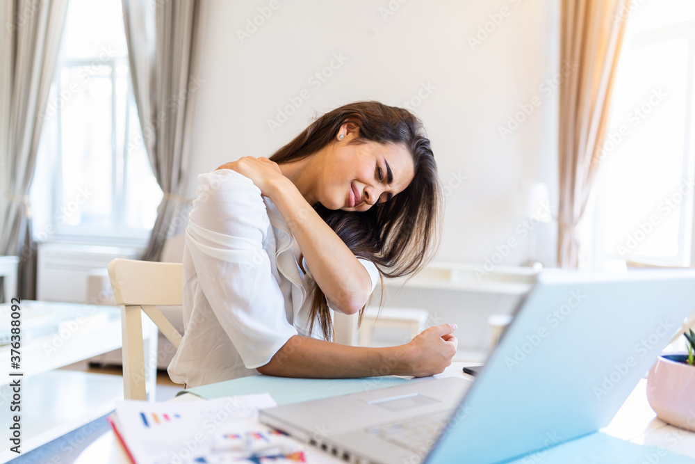 Portrait of young stressed woman sitting at home office desk in front of laptop, touching aching neck with pained expression, suffering from neck pain after working on laptop Portrait of young stressed woman sitting at home office desk in front of laptop, touching aching neck with pained expression, suffering from neck pain after working on laptop
