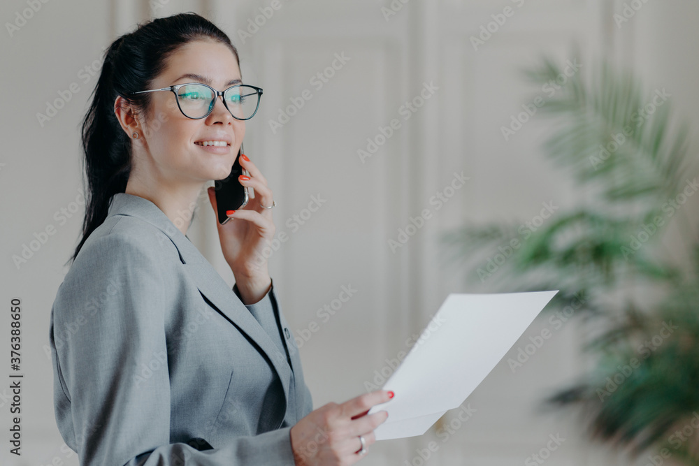 Photo of elegant brunette woman in formal clothes works with papers in office, discusses business issues via smartphone with colleague. Female manager checks information from documents, makes research Photo of elegant brunette woman in formal clothes works with papers in office, discusses business issues via smartphone with colleague. Female manager checks information from documents, makes research