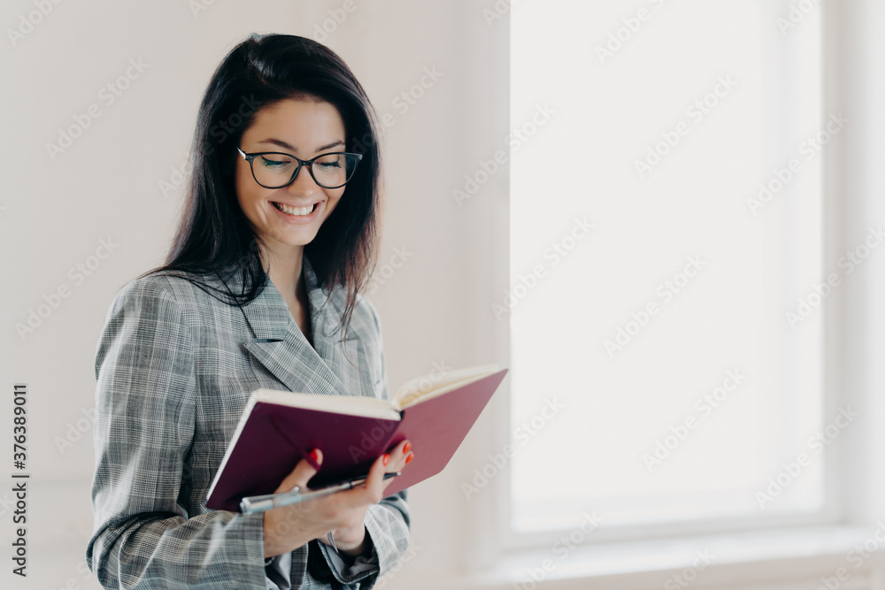 Satisfied brunette woman has happy expression writes down working plan in notepad has red manicure formal clothes records information for organization process wears spectacles notes financial schedule Satisfied brunette woman has happy expression writes down working plan in notepad has red manicure formal clothes records information for organization process wears spectacles notes financial schedule
