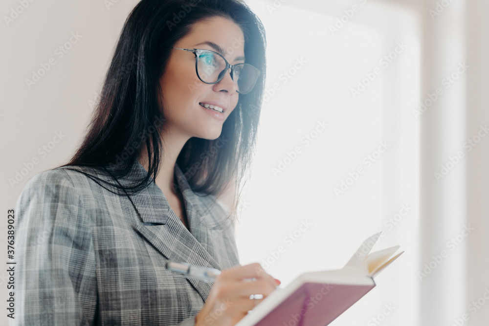 Serious thoughtful brunette European woman writes down text information in notepad, concentrated somewhere into distance, makes notes, wears formal clothes, transparent glasses, stands indoor Serious thoughtful brunette European woman writes down text information in notepad, concentrated somewhere into distance, makes notes, wears formal clothes, transparent glasses, stands indoor
