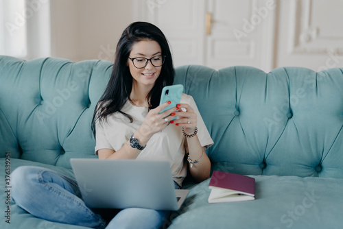 Happy female blogger chats with friends in social netwoks, works on laptop computer, poses in coworking space, sits on sofa in lotus pose, connected to wireless internet. Technology, freelancing