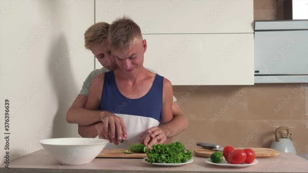 Two blond vegetarian guys hugging each other in a bright kitchen preparing breakfast. Chop vegetables for salad. Two blond vegetarian guys hugging each other in a bright kitchen preparing breakfast. Chop vegetables for salad.
