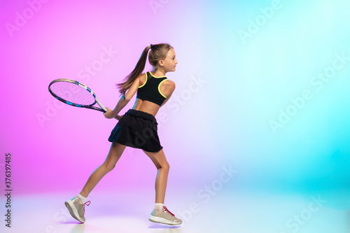 Running high. Little tennis girl in black sportwear isolated on gradient background in neon light. Little caucasian model, sport kid training in motion and action. Sport, movement, childhood concept. © master1305