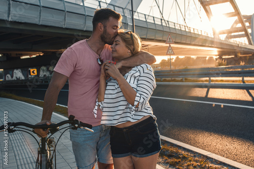 Smiling young couple with bicycle enjoying walking and embracing each other at sunset.