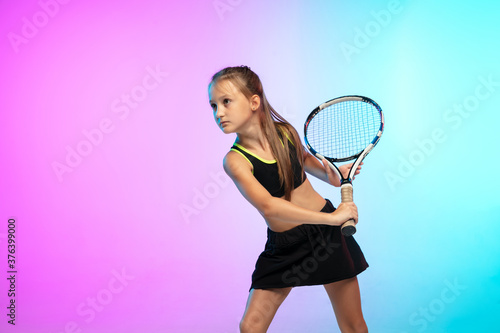 Powerful. Little tennis girl in black sportwear isolated on gradient background in neon light. Little caucasian model, sport kid training in motion and action. Sport, movement, childhood concept. © master1305