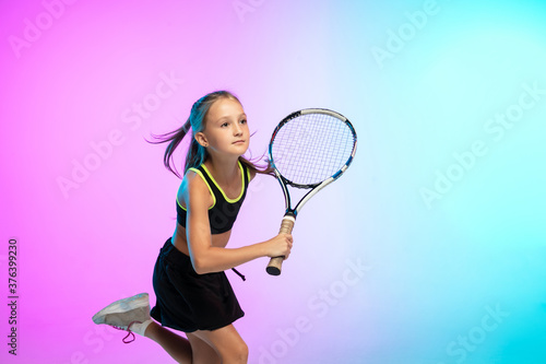 Powerful. Little tennis girl in black sportwear isolated on gradient background in neon light. Little caucasian model, sport kid training in motion and action. Sport, movement, childhood concept. © master1305