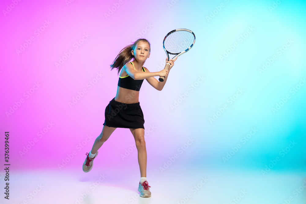 Powerful. Little tennis girl in black sportwear isolated on gradient background in neon light. Little caucasian model, sport kid training in motion and action. Sport, movement, childhood concept.