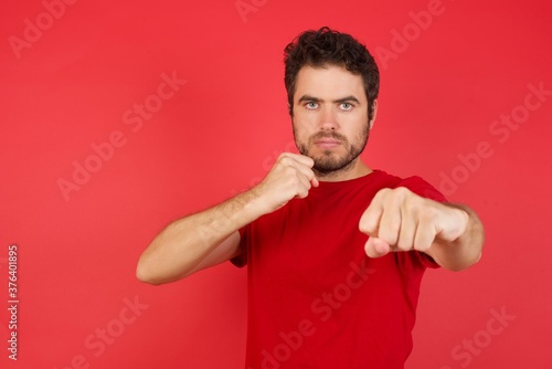 Young handsome caucasian man wearing t-shirt over isolated red background Punching fist to fight, aggressive and angry attack, threat and violence