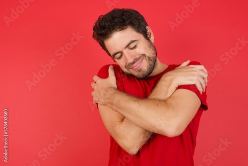 Young handsome caucasian man wearing t-shirt over isolated red background Hugging oneself happy and positive, smiling confident. Self love and self care photo