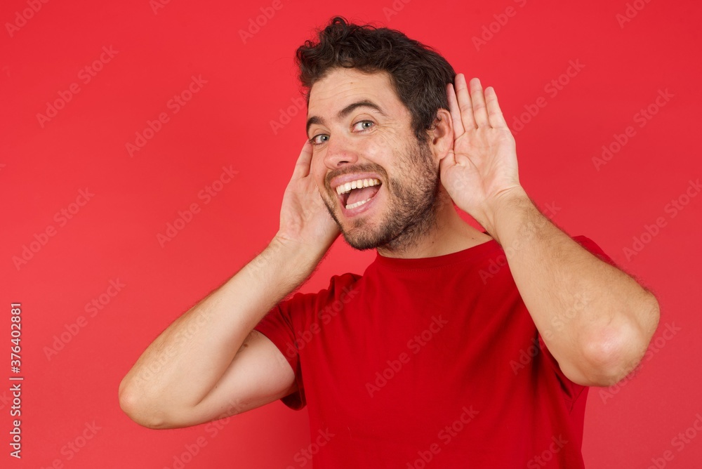 Naklejka premium Young handsome caucasian man wearing t-shirt over isolated red background Trying to hear both hands on ear gesture, curious for gossip. Hearing problem, deaf