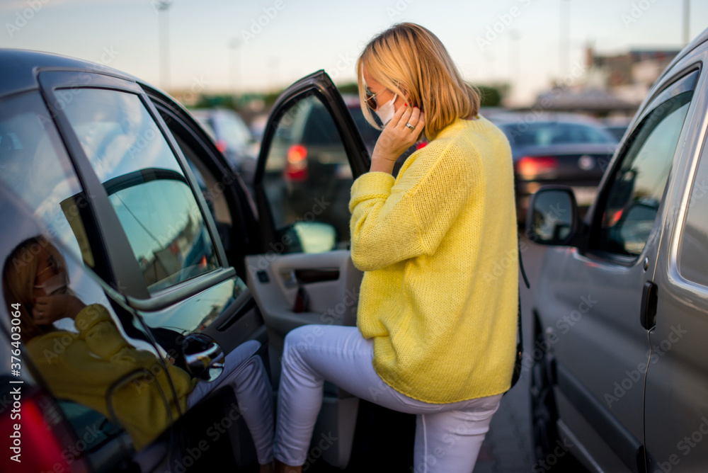 A young woman with a coronavirus mask in a yellow sweater gets into her car. The car is parked in the parking. There is bokeh in the background. Coronavirus concept. Life with covid-19. 