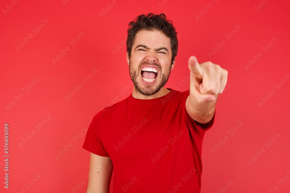 Young handsome caucasian man wearing t-shirt over isolated red background pointing displeased and frustrated to the camera, angry and furious ready to fight with you. Young handsome caucasian man wearing t-shirt over isolated red background pointing displeased and frustrated to the camera, angry and furious ready to fight with you.