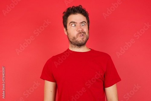 Portrait of Young handsome caucasian man wearing t-shirt over isolated red background making grimace and crazy face, screaming out of control, funny lunatic expressing freedom and wild.