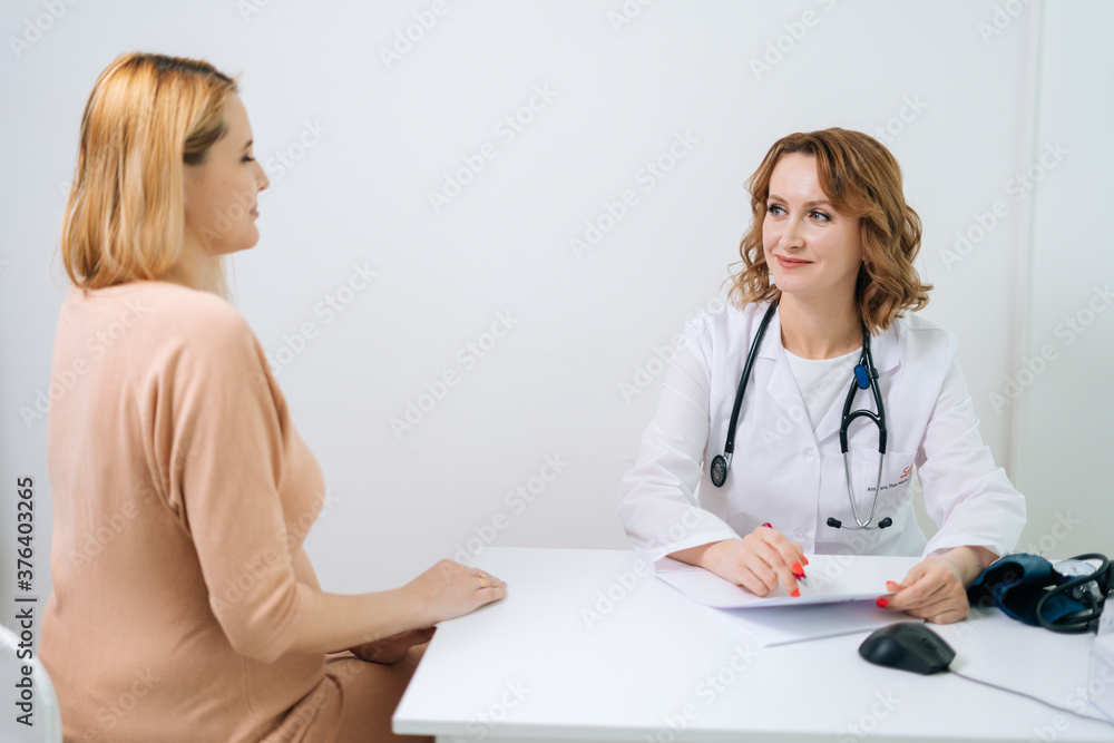 Happy young pregnant woman with blonde hair visiting doctor in prenatal clinic. Beautiful expectant mother listening doctor sitting at the desk. Concept of happy and healthy childbearing. Happy young pregnant woman with blonde hair visiting doctor in prenatal clinic. Beautiful expectant mother listening doctor sitting at the desk. Concept of happy and healthy childbearing.