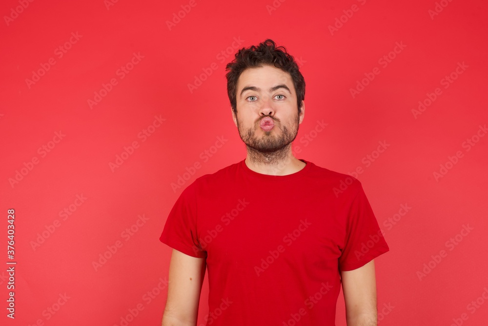 Shot of pleasant looking Young handsome caucasian man wearing t-shirt over isolated red background, pouts lips, looks with green eyes at camera, has fun with girlfriend, Human facial expressions Shot of pleasant looking Young handsome caucasian man wearing t-shirt over isolated red background, pouts lips, looks with green eyes at camera, has fun with girlfriend, Human facial expressions