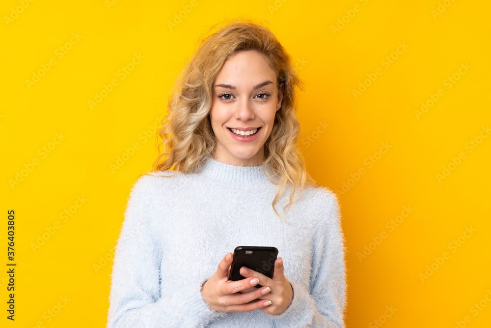 Young blonde woman isolated on yellow background sending a message with the mobile Young blonde woman isolated on yellow background sending a message with the mobile
